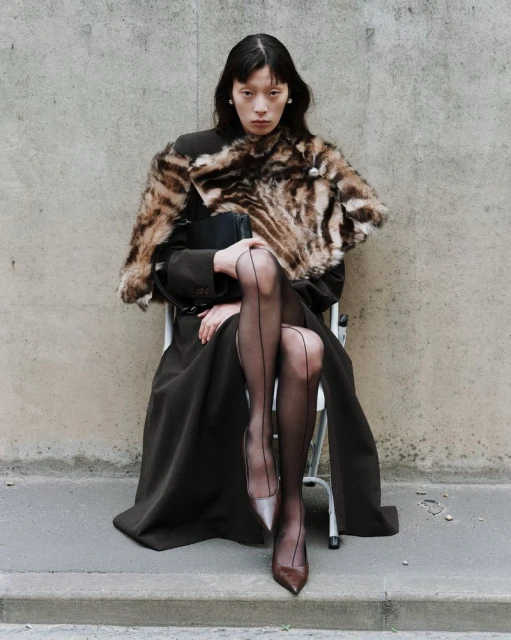 Elegant woman poses seated wearing fur stole, dark coat, sheer hose, and pumps against a concrete wall in an outdoor urban environment.