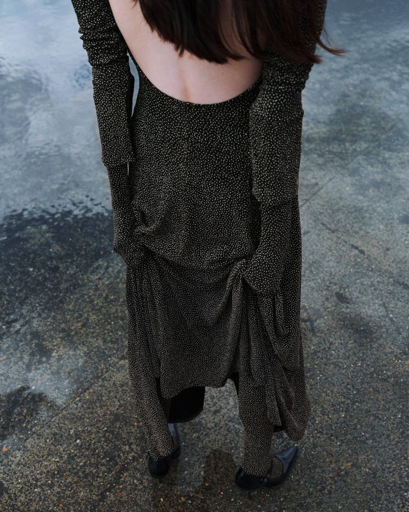 An adult woman with her back to the viewer wears a long sleeve, open-back black dress with gold flecks standing on a mottled gray surface.