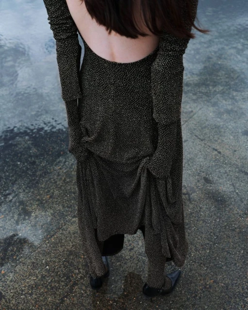 An adult woman with her back to the viewer wears a long sleeve, open-back black dress with gold flecks standing on a mottled gray surface.