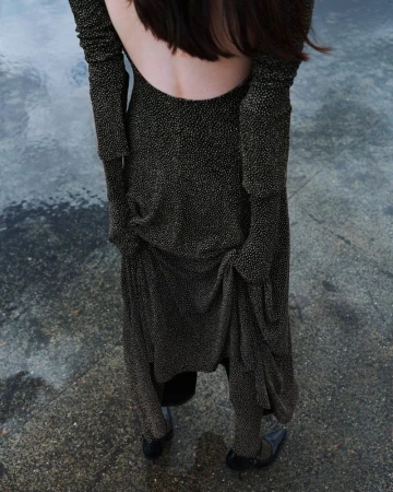 An adult woman with her back to the viewer wears a long sleeve, open-back black dress with gold flecks standing on a mottled gray surface.