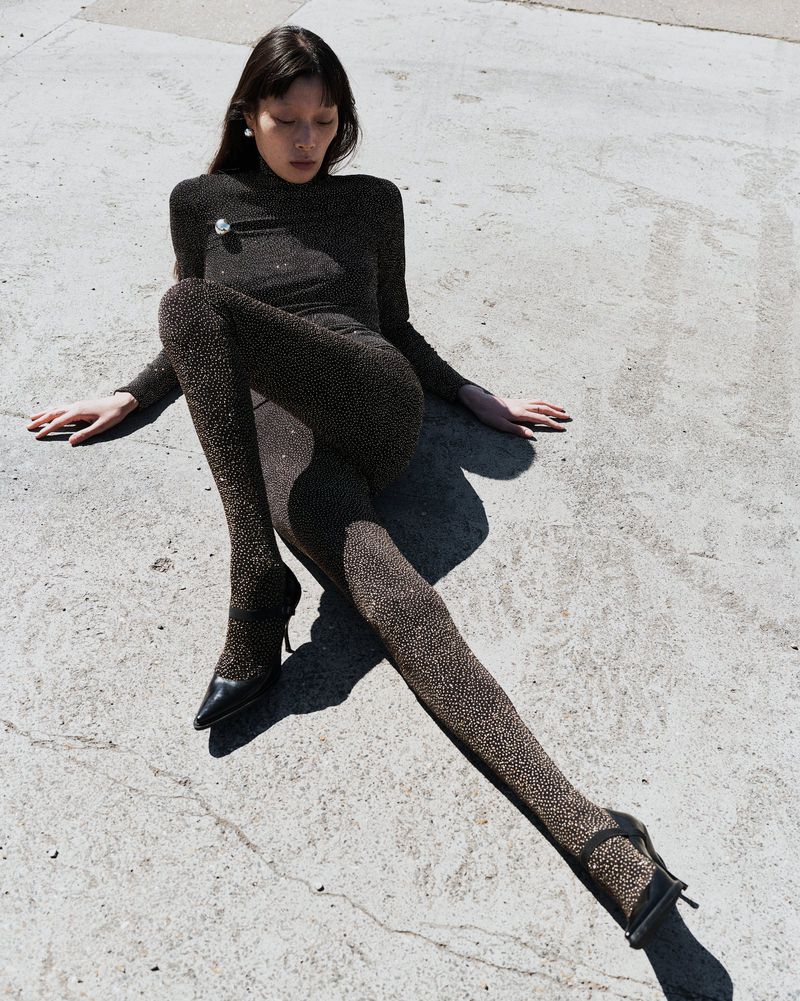 Model Jay Pak in a sparkling dark catsuit seated outdoors on grey concrete pavement for Beaufille Resort 25