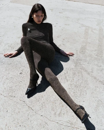 Model Jay Pak in a sparkling dark catsuit seated outdoors on grey concrete pavement for Beaufille Resort 25