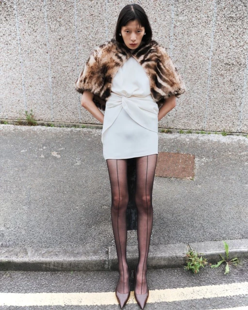 An Asian woman models a white dress, fur stole, and sheer black tights outdoors. The look is captured in a medium shot against a concrete wall.