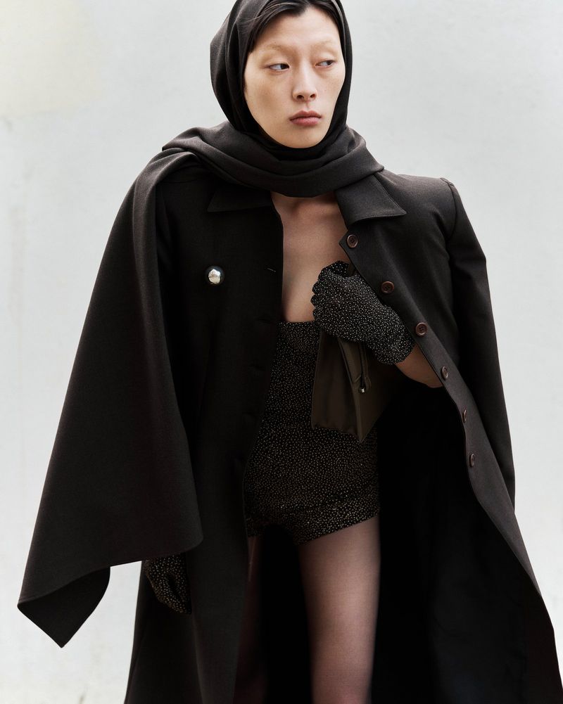 Elegant adult woman in a hooded scarf and cape, posing indoors against a white backdrop, showcasing dark fashion with soft, diffused lighting.