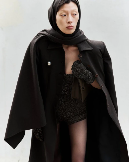 Elegant adult woman in a hooded scarf and cape, posing indoors against a white backdrop, showcasing dark fashion with soft, diffused lighting.