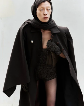 Elegant adult woman in a hooded scarf and cape, posing indoors against a white backdrop, showcasing dark fashion with soft, diffused lighting.