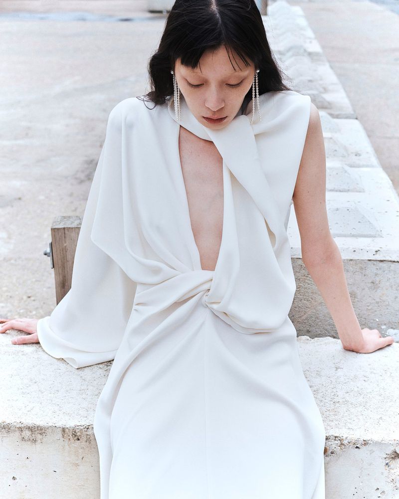 Elegant adult woman in a flowing white dress with a deep neckline, seated on concrete steps, showcasing fashion and minimalist style outdoors.