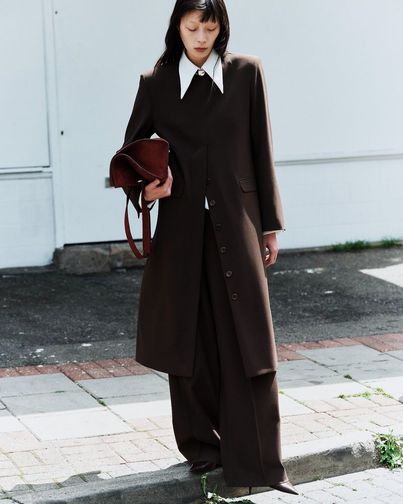 Model standing on a sidewalk wearing a dark brown long coat and matching trousers while holding a reddish brown handbag