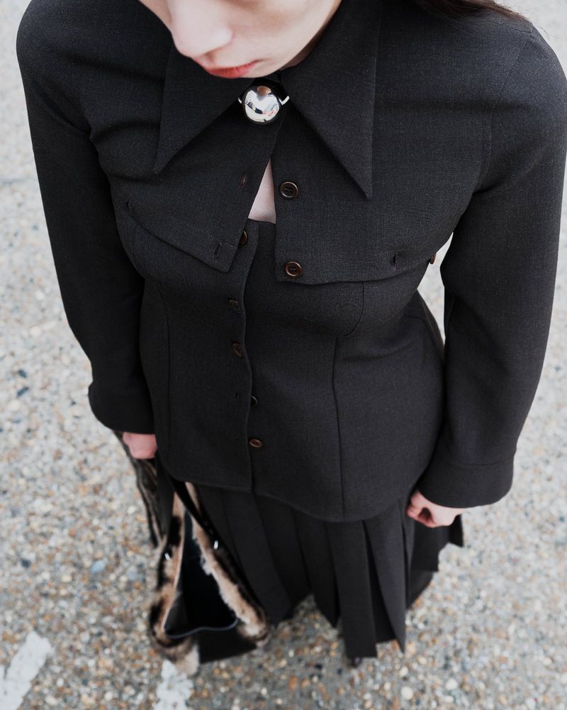 High-angle shot of a young woman in a dark jacket and pleated skirt with a vintage purse on pavement, showcasing modern fashion.