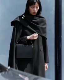 Model wearing a black coat and cape holding a black handbag against an outdoor textured background