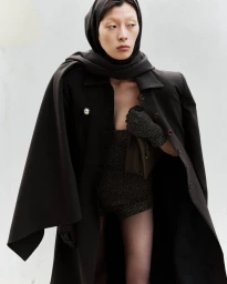 Model Jay Pak wearing a dark coat and matching headscarf against a plain white background for Beaufille Resort 25