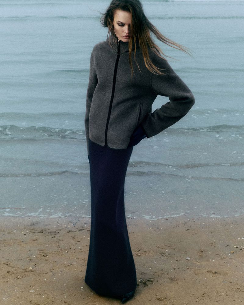 A woman in a gray jacket and long black skirt poses on a sandy beach with a hazy ocean backdrop under overcast lighting conditions.