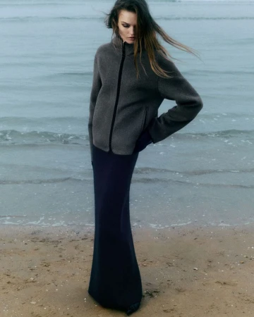 A woman in a gray jacket and long black skirt poses on a sandy beach with a hazy ocean backdrop under overcast lighting conditions.