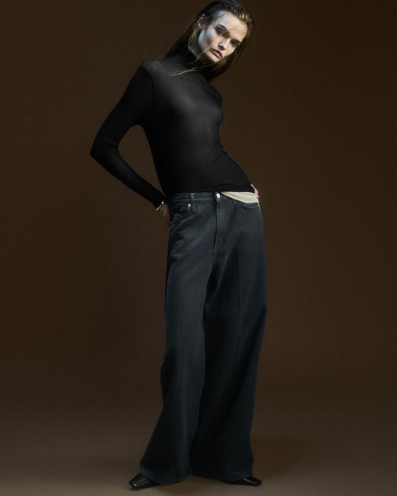Woman poses in black turtleneck and baggy jeans against a brown backdrop in studio lighting. Fashion editorial shot, full body view.