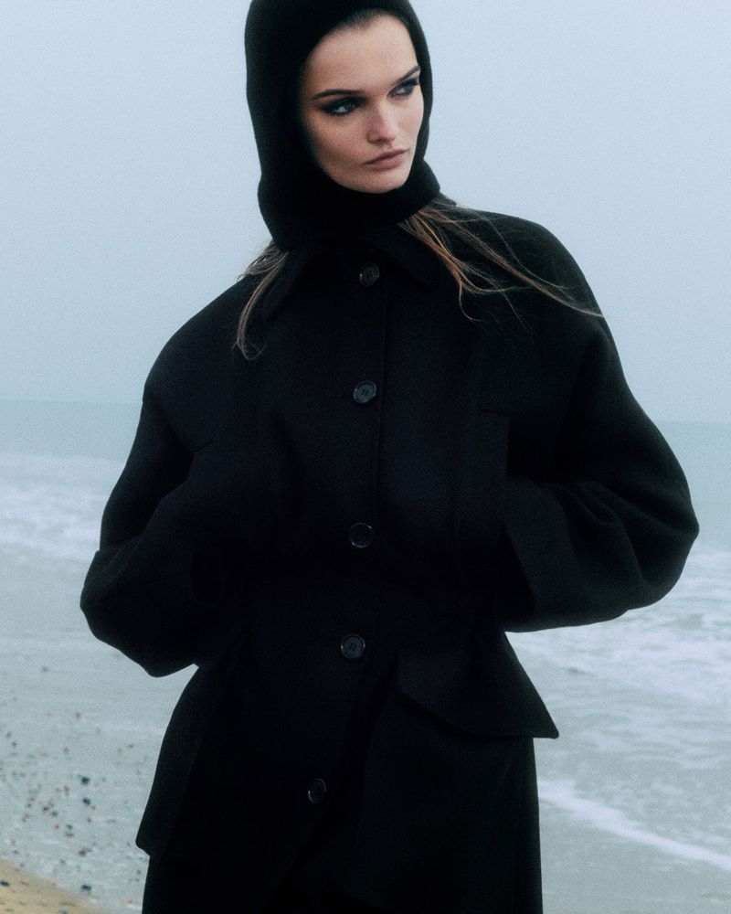 A fashionable adult woman poses in a black hooded jacket on a misty beach. The image emphasizes the jacket's design in an outdoor setting.