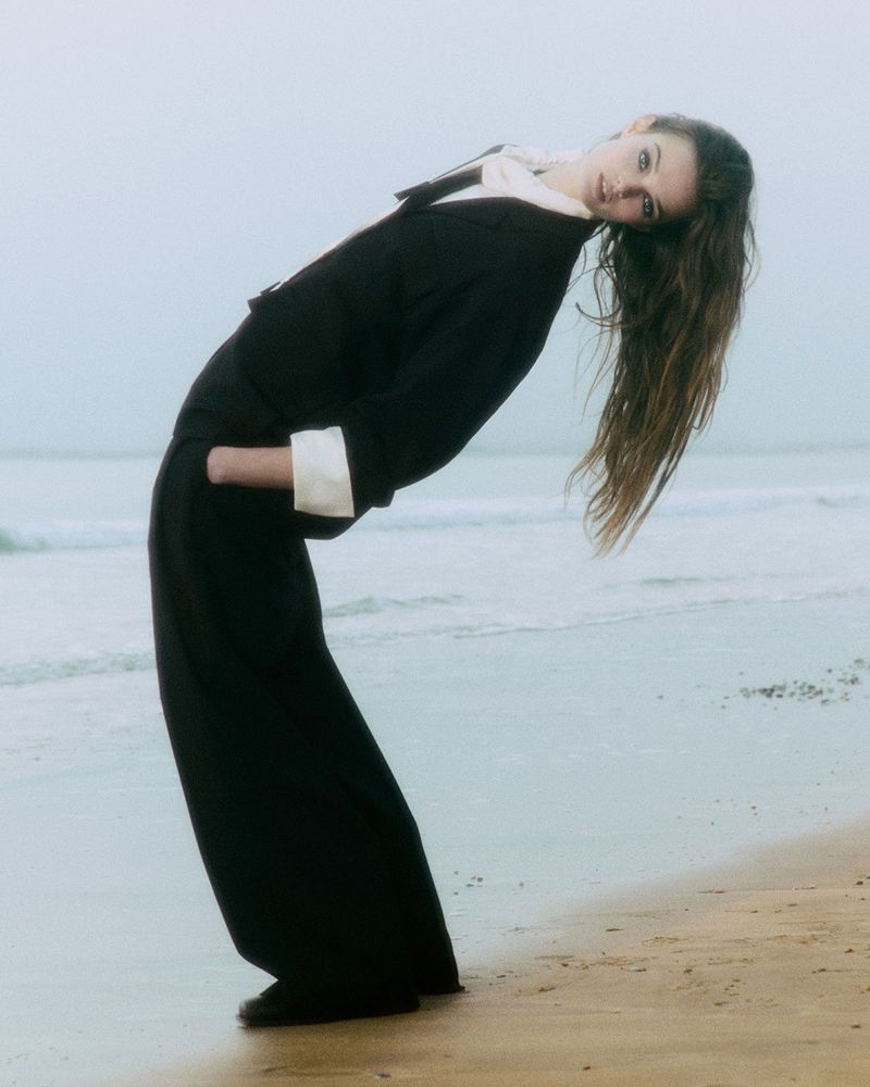 Fashion shot of a woman in black wide-legged pants and a black jacket with white collar and cuffs, standing on a sandy beach with ocean backdrop.