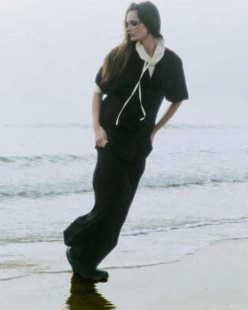 Model Lulu Tenney in dark clothing posing on a sandy beach for COS 2025 Collection, photographed by Daniel P. Shea.