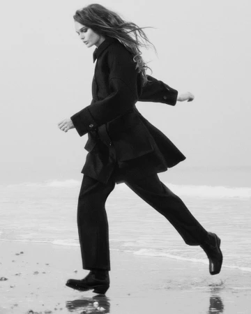 Model Lulu Tenney running on a sandy beach wearing a dark COS coat and trousers for the 2025 Collection.