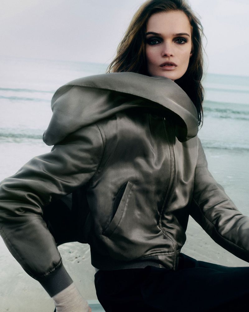 A woman in a stylish grey hooded jacket poses on a beach with a neutral expression, with ocean waves in the background, shot at eye-level.