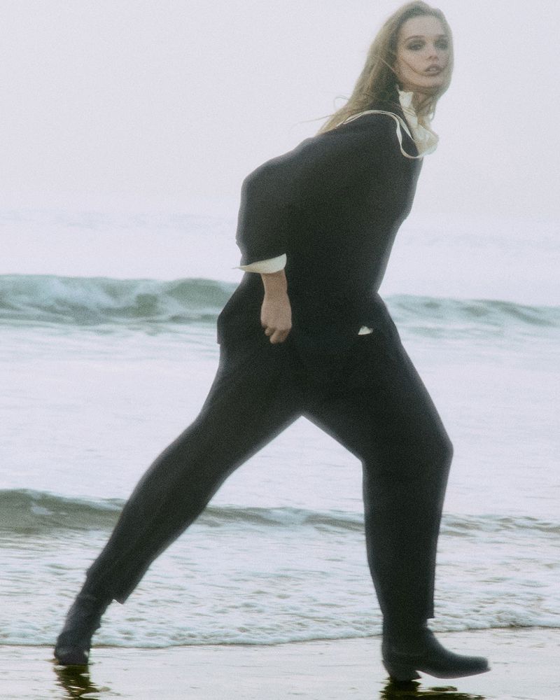 A woman in black walks on a beach in a wide shot, wearing a dark top and pants with waves and sky in the background, fashion.