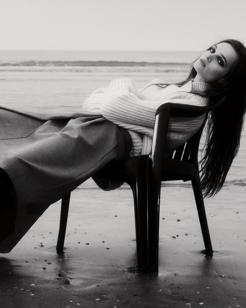 Black and white portrait of a young adult woman relaxing on a plastic chair on a beach with ocean waves in the background.