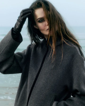 Model Lulu Tenney wearing a dark grey coat and leather gloves with an ocean background for COS 2025 collection.