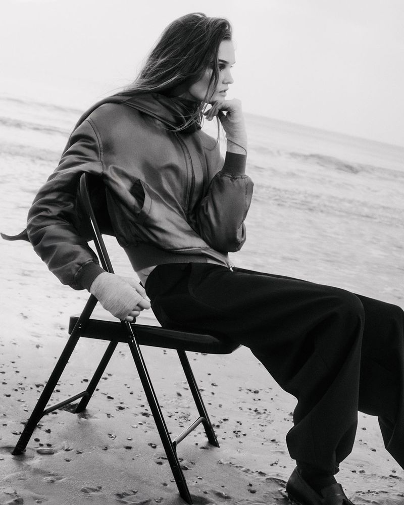 A young woman in a jacket and pants sits on a black folding chair at the beach, with the ocean and sky visible in the background.