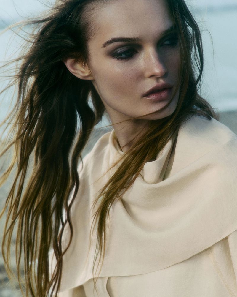 Woman with windswept brown hair and light complexion wears a cream-colored top in an outdoor setting, near a sandy beach.