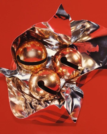 Three golden jingle bells are nestled inside shiny, reflective paper wrapping against a solid red background, creating a festive scene.