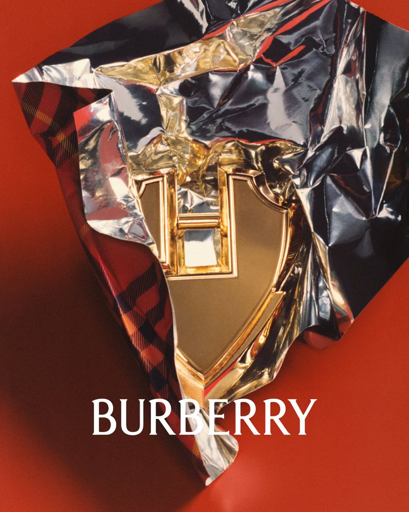 Burberry logo displayed in gold metallic with tartan fabric and silver foil on a solid red background in a product shot.