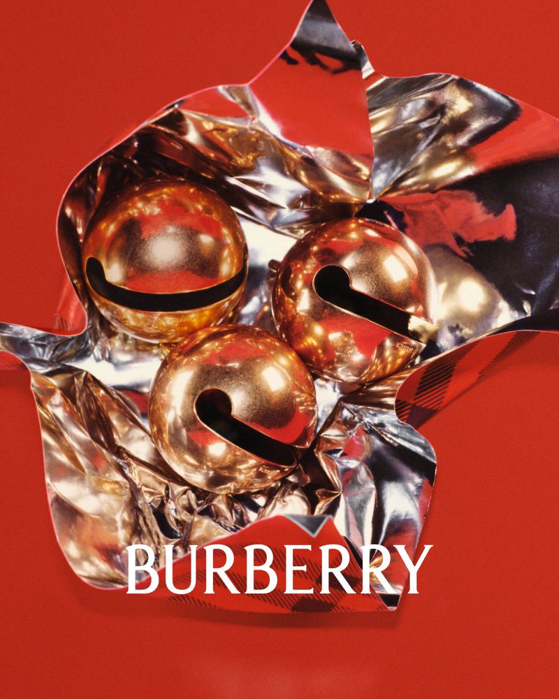 Three golden jingle bells wrapped in silver and red shiny packaging, featuring Burberry branding on a red background.