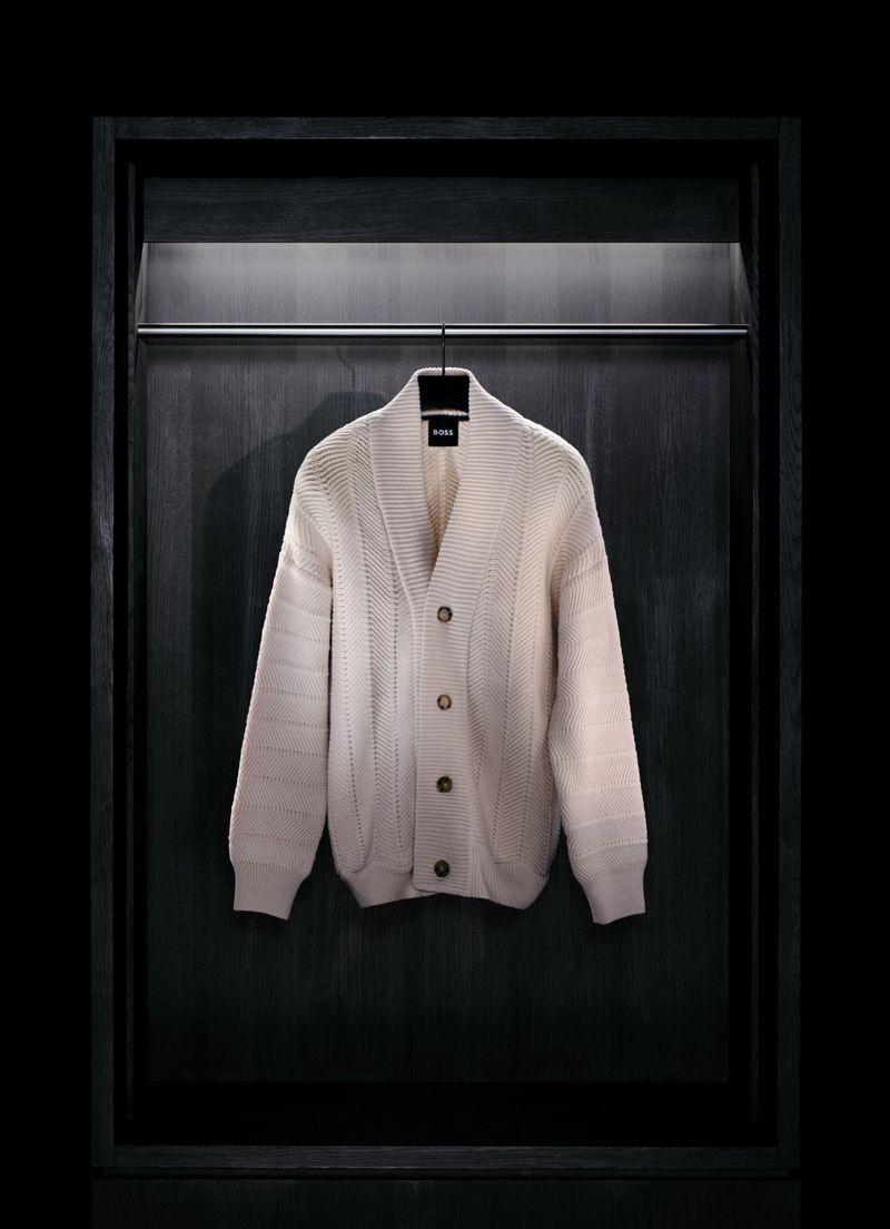 Cream-colored knit cardigan with buttons hanging on a metal rack against a dark paneled background, in a minimalist fashion still life.