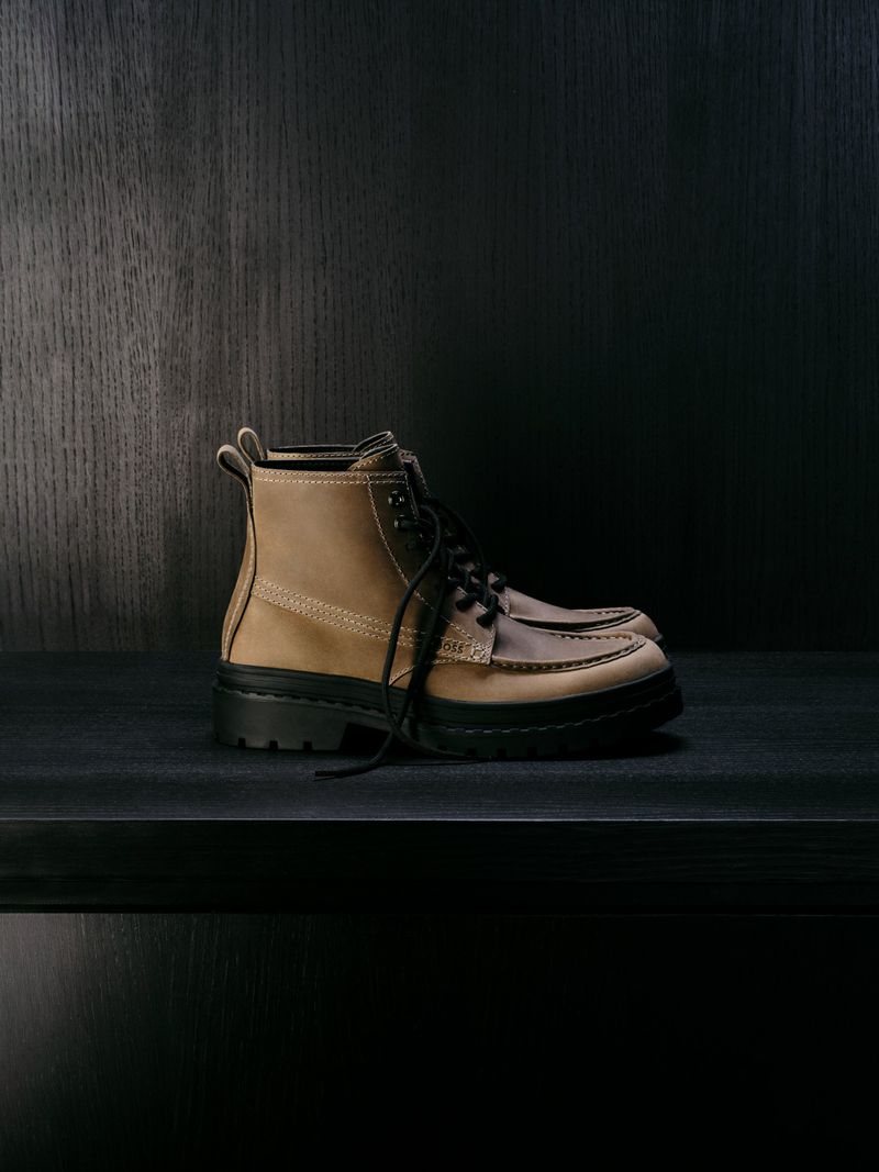 Tan and brown leather work boots with black laces sit on a dark shelf against a dark wood paneled wall in a product shot.
