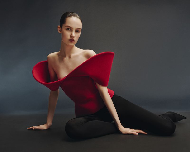 Model seated on a dark studio floor wearing a structured red top with flared shoulders and black trousers