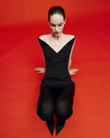 Adult female in a sleek black dress sits on a vibrant red surface, gazing downward in a studio setting, presenting a minimalist fashion editorial.