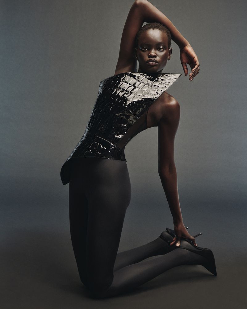 Fashion editorial shot of a Black female model kneeling in a high-fashion shiny black corset top and dark tights in a studio.