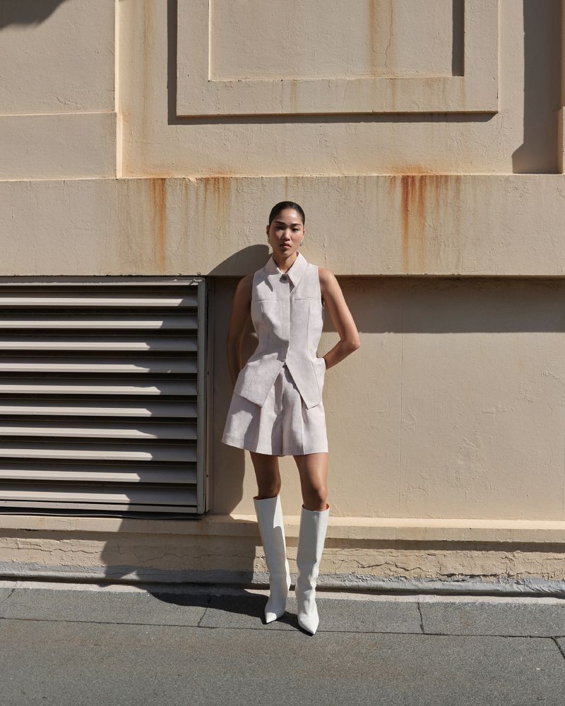 A woman poses outdoors wearing a pale linen vest and matching shorts, paired with white knee-high boots, against a neutral wall.