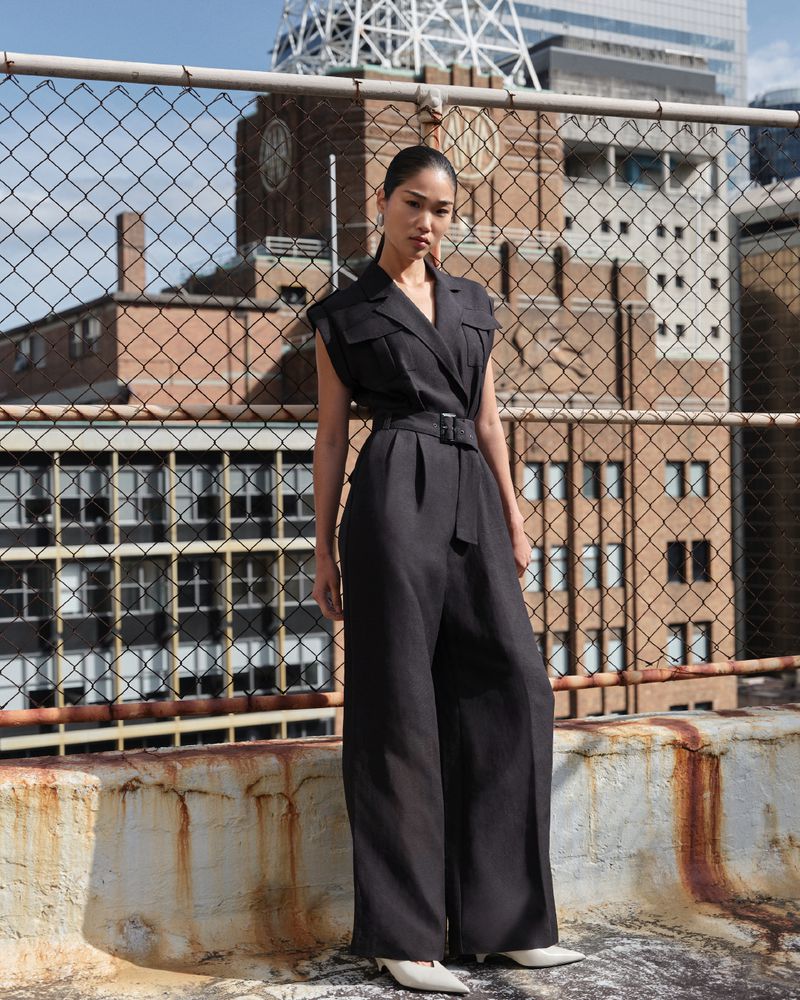 A slender Asian woman in a dark sleeveless jumpsuit poses on a rooftop with a chain-link fence, set against a backdrop of brick buildings and skyscrapers.
