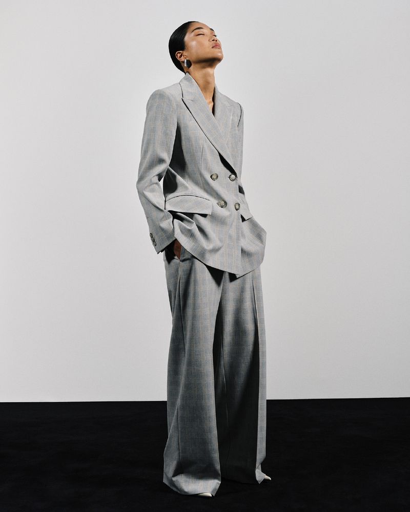 An Asian woman poses in a gray double-breasted suit with wide-legged pants, looking up in a brightly lit photo studio against a gradient background.
