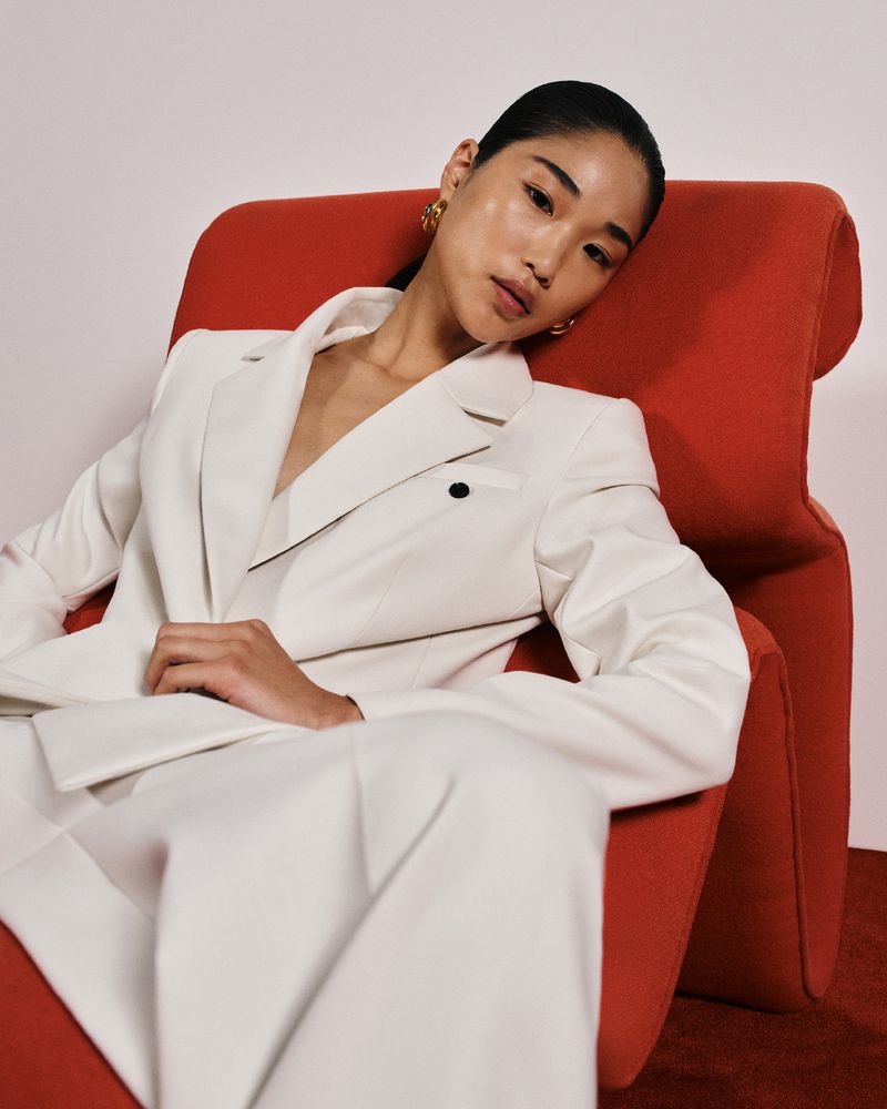 Model in white suit jacket seated on a bright red armchair against a neutral background