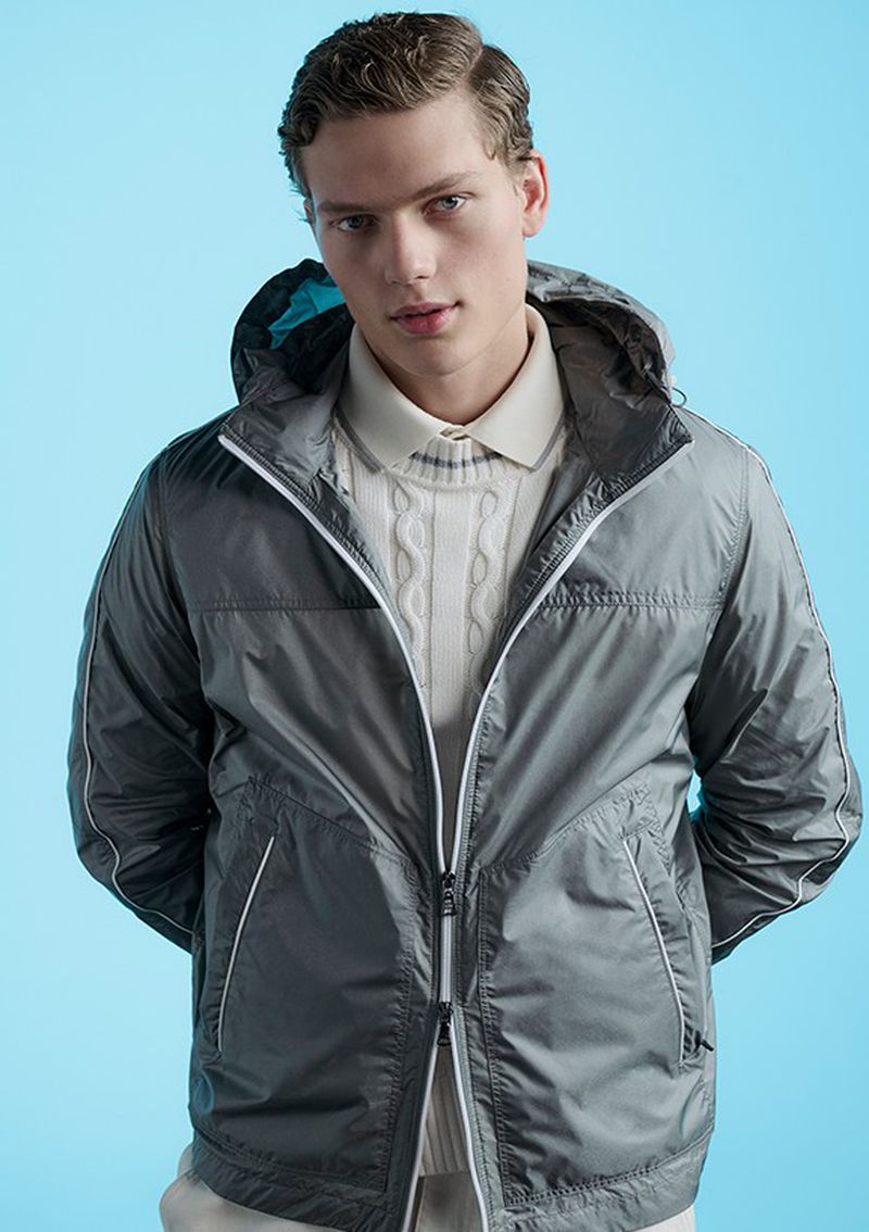 Male fashion model poses in gray hooded jacket and cream cable knit sweater against a light blue studio backdrop, showcasing modern menswear.