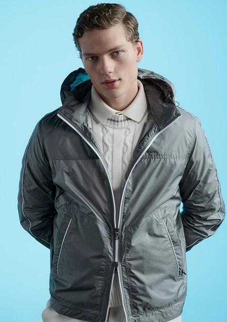 Male fashion model poses in gray hooded jacket and cream cable knit sweater against a light blue studio backdrop, showcasing modern menswear.