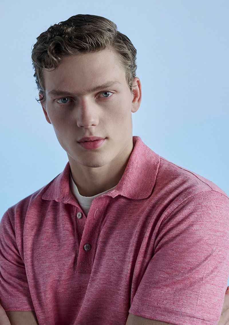 Headshot of a young adult male with light skin, wavy light brown hair, and blue eyes wearing a heathered red polo shirt in a studio.