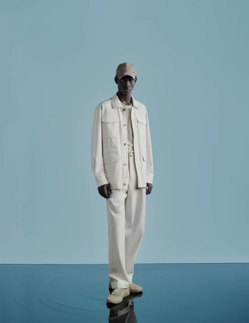 Full-length shot of a Black male model in light beige casual wear, including a jacket, cap, and sneakers, against a pale blue backdrop.
