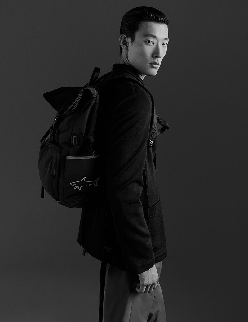 A portrait of an Asian male model wearing a black blazer and carrying a black backpack, shot in a photo studio with dark background.