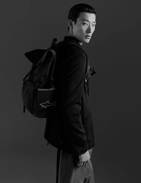 A portrait of an Asian male model wearing a black blazer and carrying a black backpack, shot in a photo studio with dark background.