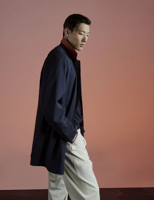Asian man in a navy overcoat poses indoors against a gradient coral background, with his hands in his pockets and wearing cream trousers.