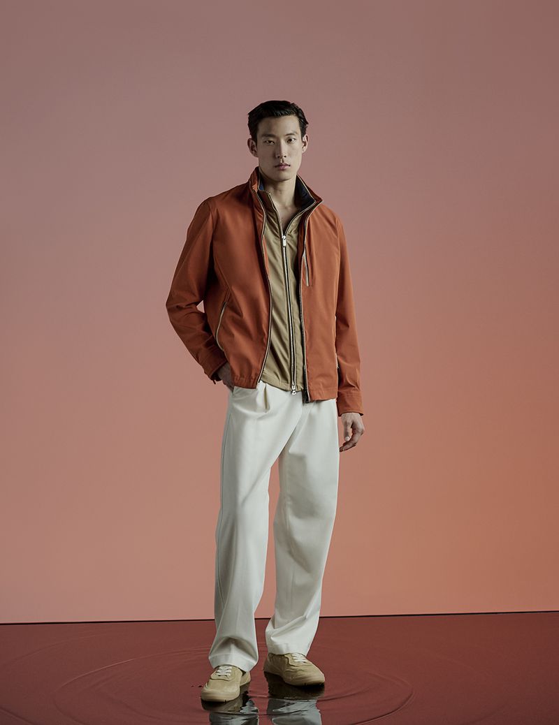 Portrait of a male model in a rust-colored jacket and white trousers against a neutral backdrop, showcasing modern menswear.