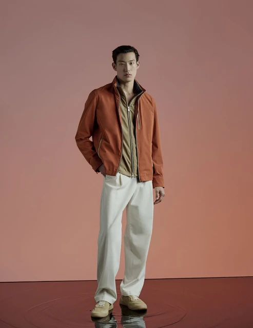Portrait of a male model in a rust-colored jacket and white trousers against a neutral backdrop, showcasing modern menswear.