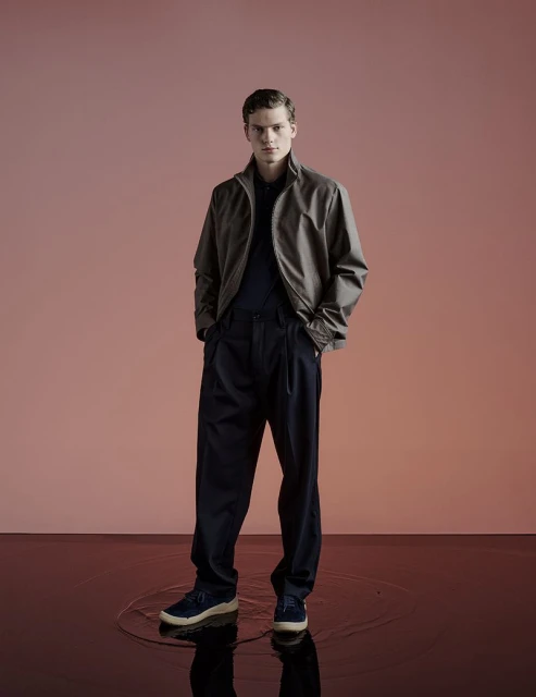 Full-length shot of an adult male model in a taupe jacket, dark blue pants and matching sneakers against a gradient pink backdrop.
